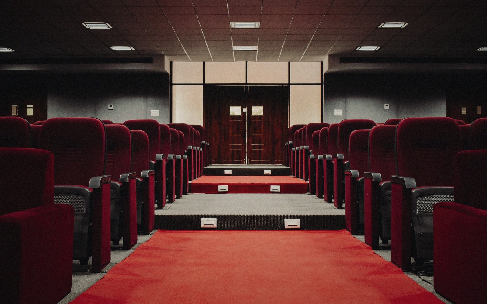 Empty theater aisle lined with red velvet seats leading to wooden double doors